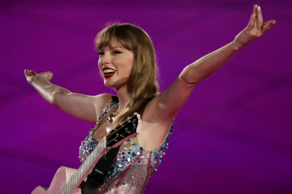 Taylor Swift performs with a guitar, wearing a sequined outfit. Arms wide open