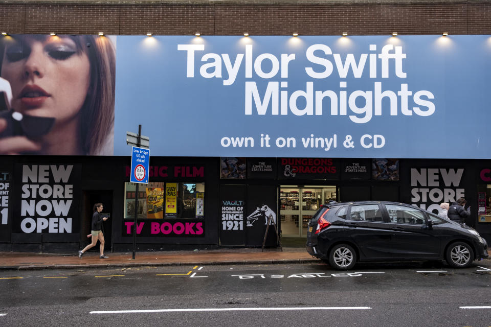 Billboard featuring Taylor Swift advertising her "Midnights" album available on vinyl and CD above a street scene with shops and a car