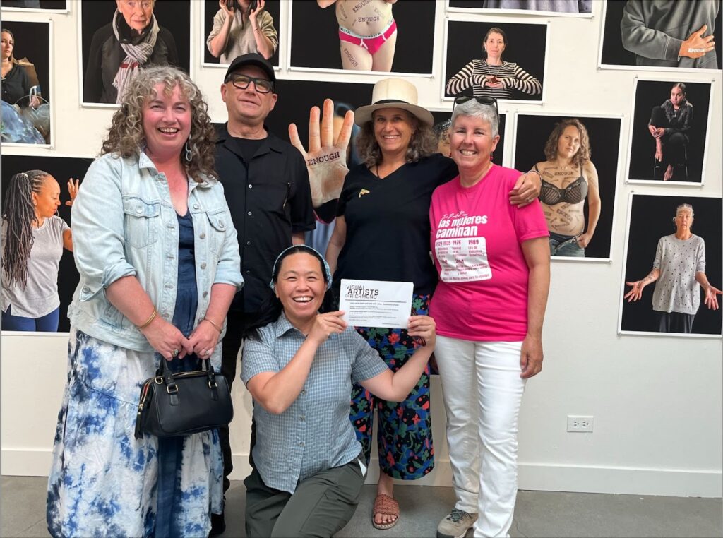 From left, Erin McLuskey Wheeler in jean jacket and blue and white long skirt clutching a black handbag; Brian Conery dressed in black with black cap and black rimmed glasses; Lauren Ari, kneeling with a blue button-down shirt and gray pants, a blue hairband over dark hair and holding up a sheet of paper that says something about Visual Artists of Richmond; Rebeca Garcia-Gonzalez in a white tophat, black top and floral skirt, holding a giant hand with the word 'enough' written across it, and Irene Wibawa wearing a hot pink T-shirt and white pants, with short white hair. They all are standing in front of a wall of various paintings.