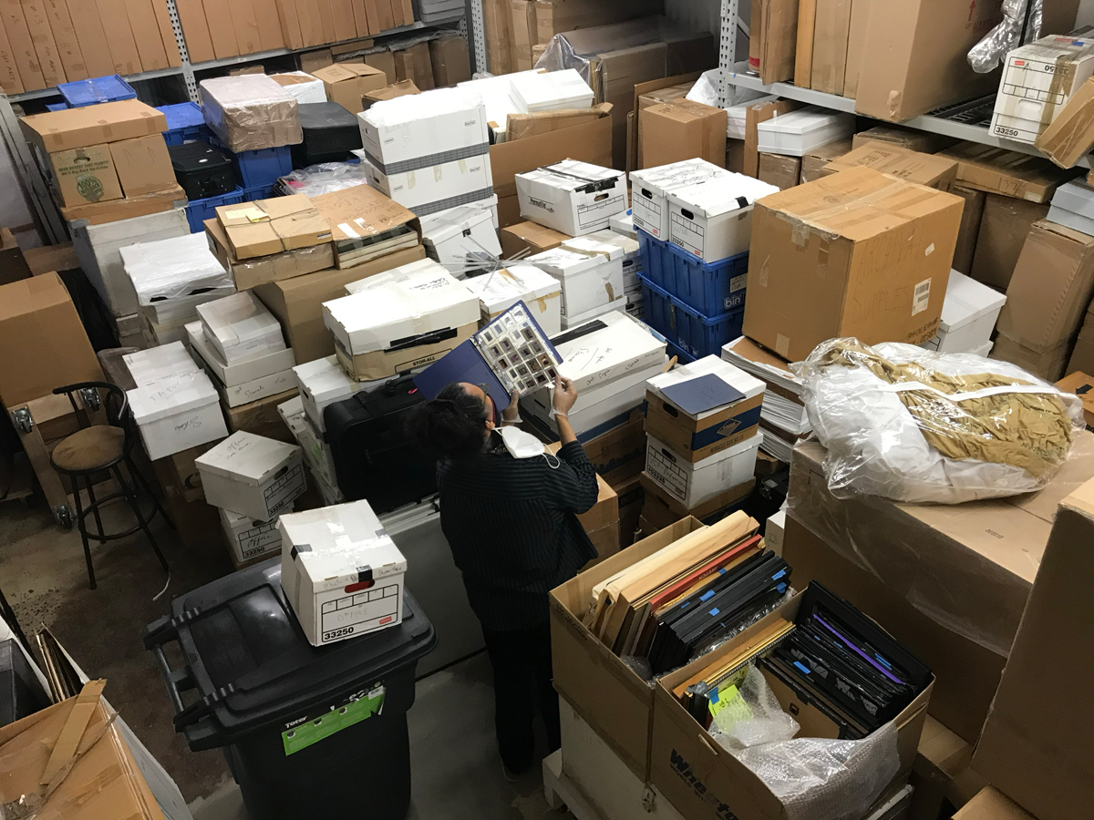 A Black man seen from behind looks at a binder of slides of artwork. He is surrounded by dozens of boxes.  