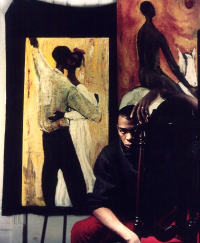 Geoffrey Holder, a young Black man sits in a chair backward, with two paintings behind him. 