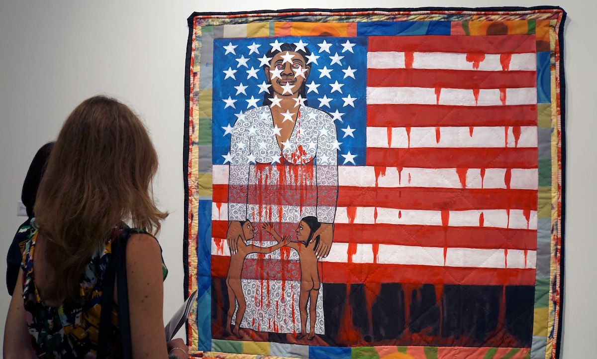 A woman looking at a painting of an American flag inset with a Black woman holding two Black children at her feet. Both the woman and the flag appear to bleed.