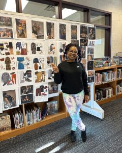 Artist Jasenya McCauley can be seen standing in front of the large bulletin board she brought in for her presentation. She is smiling widely and holding up a peace sign. Behind her on the board are rows and rows of outfits she had curated for clients- these outfits appear on TV and occasionally the theatre.