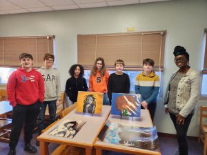 Seven people are seen standing in ront of a table with four mixed- media art pieces displayed on top. All of these individuals are Sand Creek students except for artist D. Colin- seen on the far right of the picture. All are smiling for the camera.