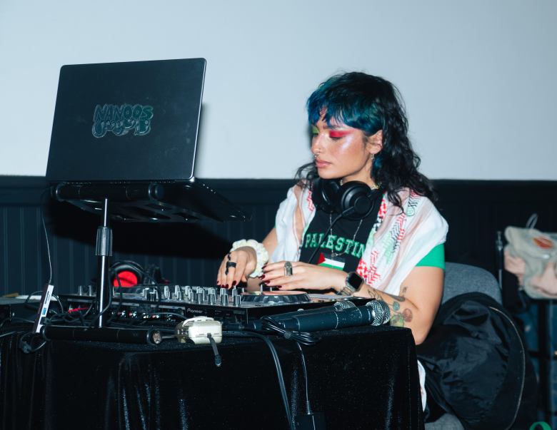 A portrait of a person DJing behind a mixer and laptop setup. They have on bright red eyeshadow, a t-shirt that says Palestine and a colorful kaffiyeh. They are looking down at the mixer.