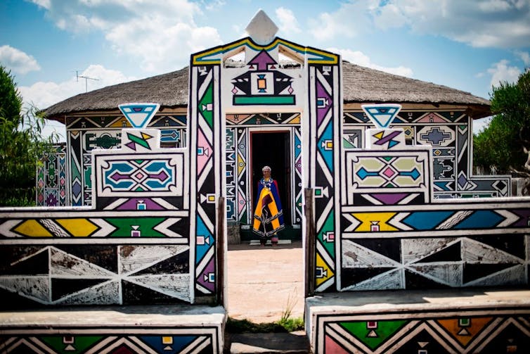 A woman stands in beaded attire in the entrance to a house which has geometric patterns painted on it.