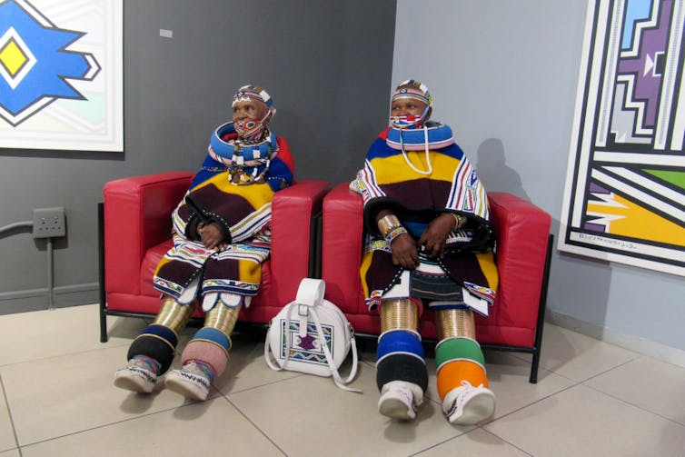 Two women sit in red chairs wearing extraordinary outfits of beaded blankets, outsize beaded necklaces, bracelets down the legs and beaded bottoms of the legs.