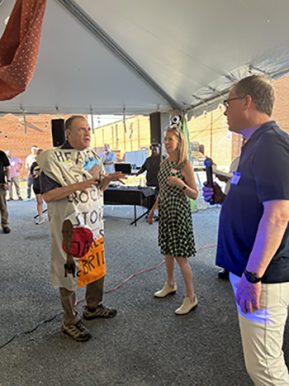 A man wearing a sack talks to a woman and another man who is holding a microphone. 