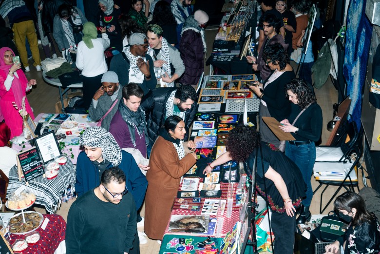 An overhead shot of an Artists Against Apartheid market. The room is filled with vendors at tables and people looking at the displays. Several wear kaffiyehs.