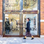 A person walks by a brick building with glass doors that have a logo that reads New York with arrows.