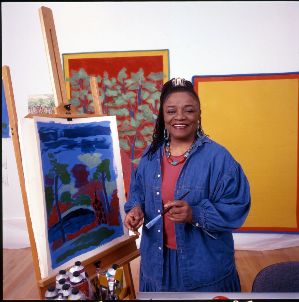 A Black woman in a long denim shirt smiling beside a painting of a landscape on an easel.