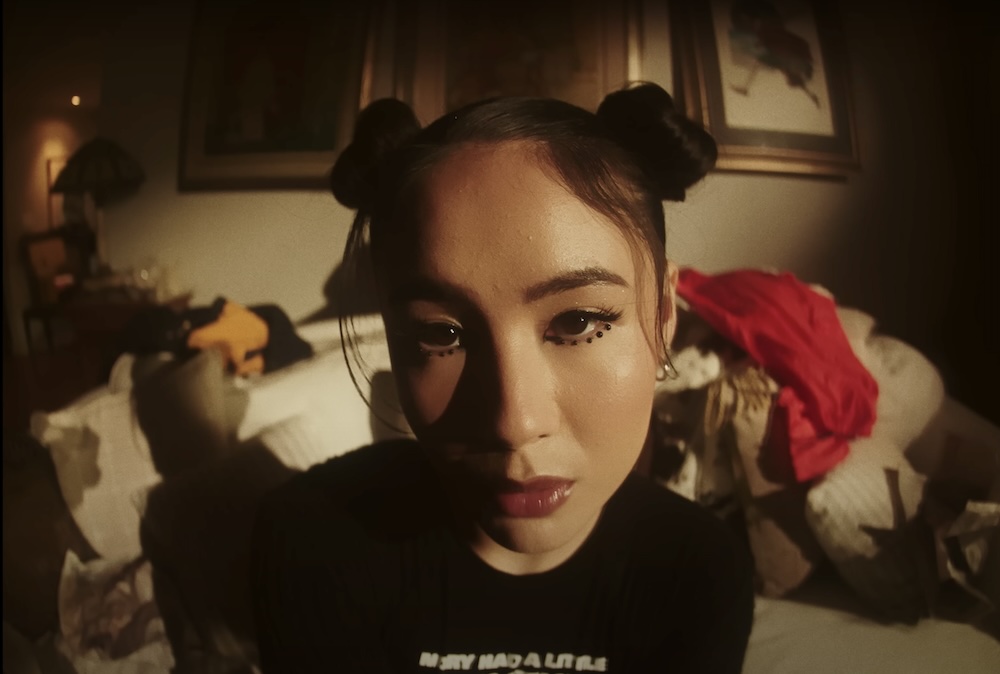 Artist Emei poses in front of camera with fish eye lens, wearing space buns and a black t shirt