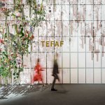 Two blurred figures walk past a wall that says TEFAF. At left is a suspended installation containing various flowers and other plants.