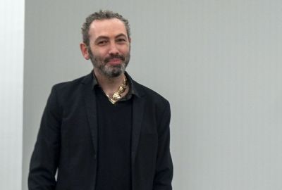 A man in a black shirt and a black suit jacket smiling.