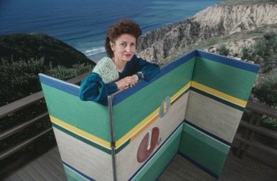 A white woman with brown hair behind a green folding screen against a backdrop of a seascape.