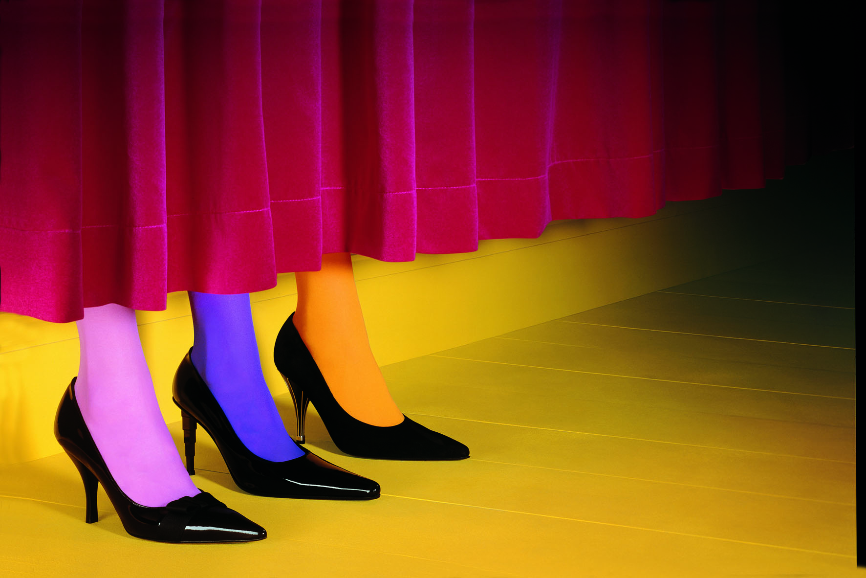 Three feet wearing black heels and colorful tights emerge from behind a red curtain