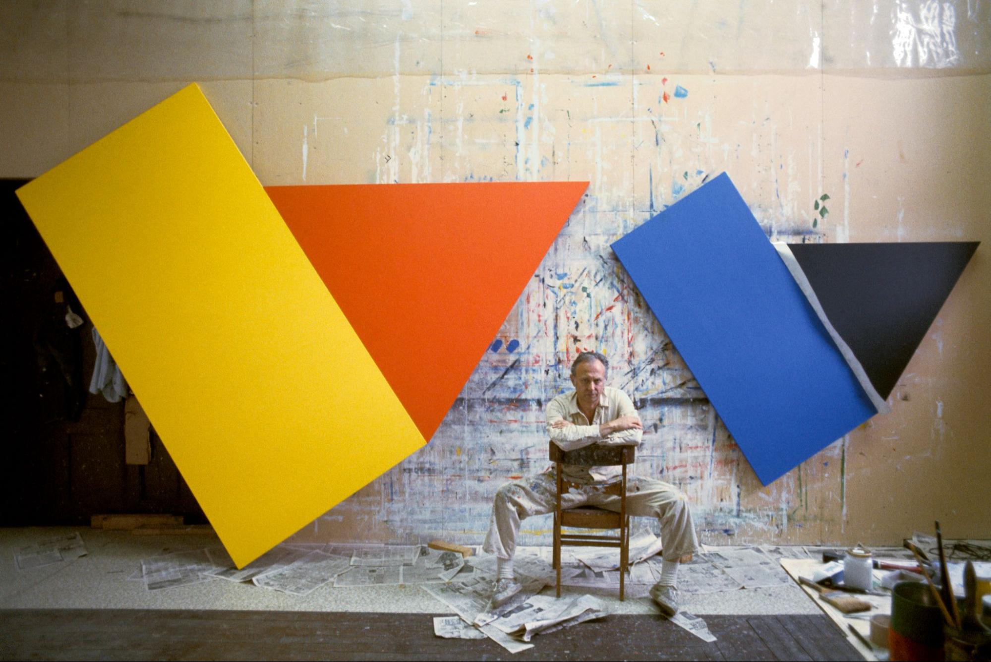 A man sits in a chair in front of an artwork made of colorful shapes