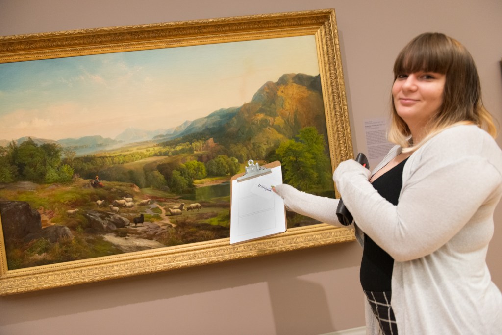 A teacher participating in a workshop holds up a card that reads tranquil and gestures towards the landscape behind her.