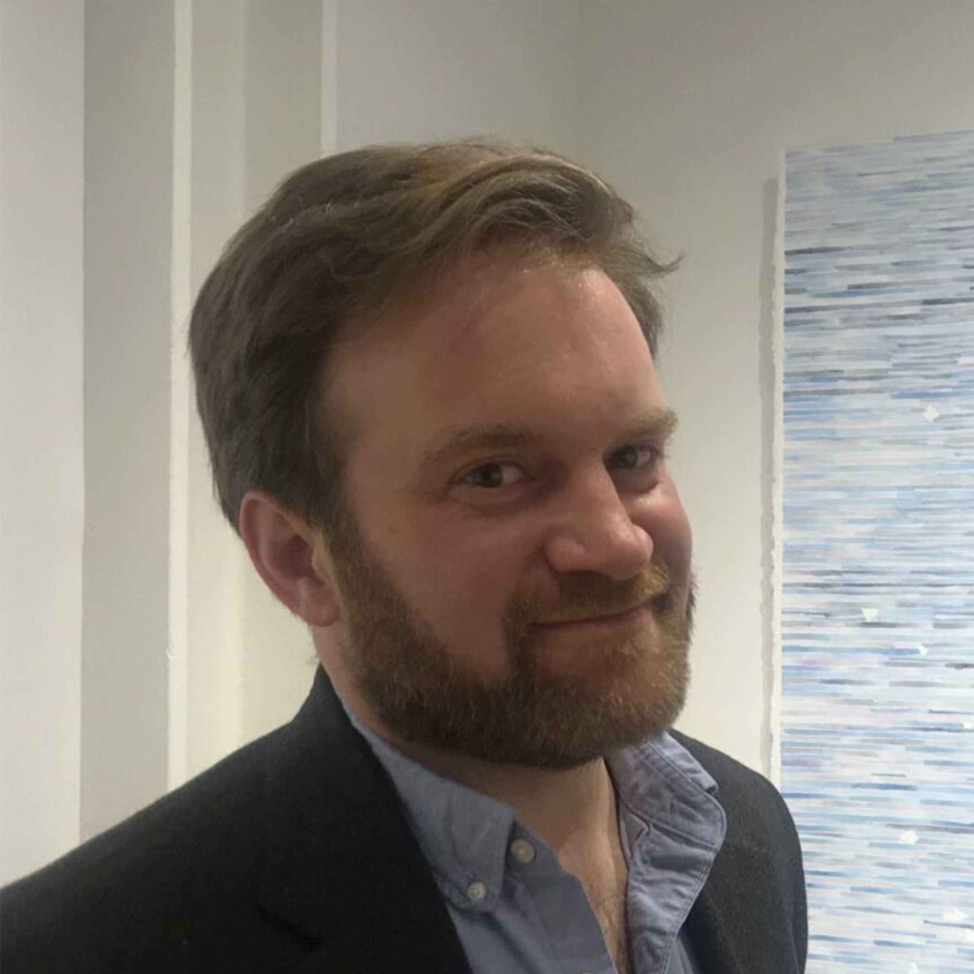 Photo: A headshot of a man with light brown hair and a beard. He has a half smile and is wearing a nice suit