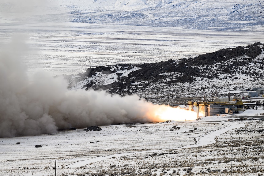 A wide shot of ball of fire and smoke firing out of a white building. In the back are snow-covered hills.