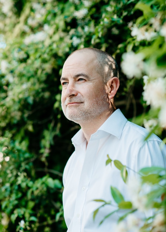 A middle-aged man with shaved grey hair and a white shirt is portrayed from his left-hand profile against a leafy green, blurry background as he stares into the distance.