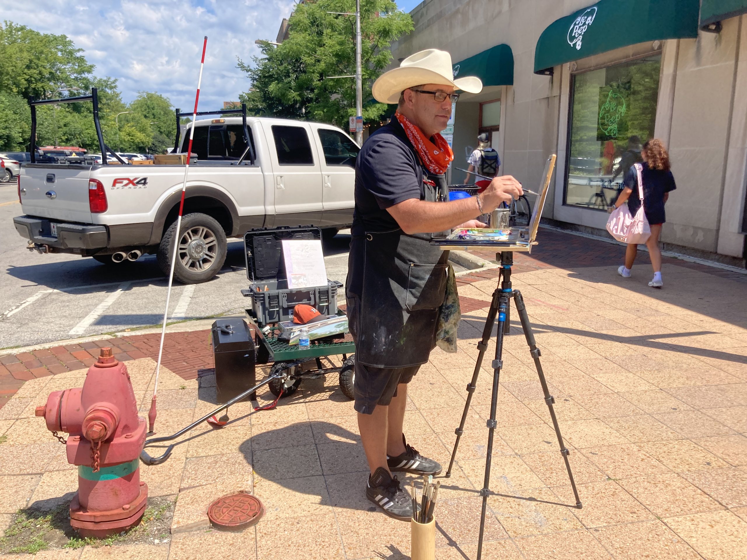 man in cowboy hat set up, standing and painting on sidewalk