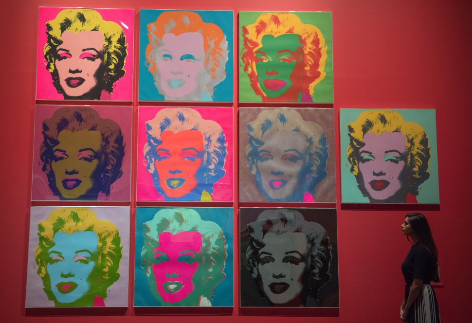 A woman stands in front of a red wall that has 10 square portraits of Marilyn Monroe, painted by pop artist Andy Warhol.