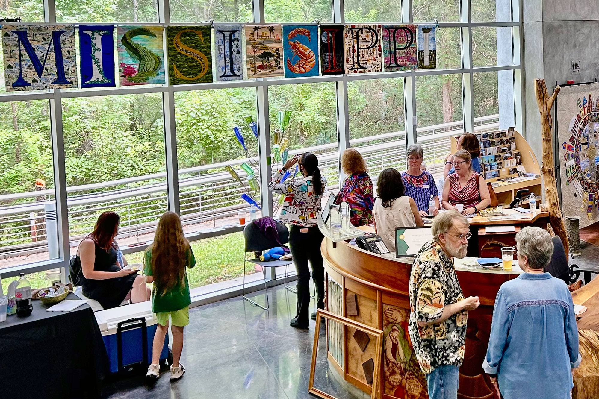 A crowd is seen in a large room lit with natural light through a glass window wall. It's decorated with art that spells out Mississippi