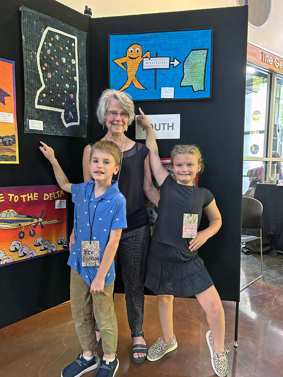 A woman and two children point to a green quilted art piee depicting Mississippi