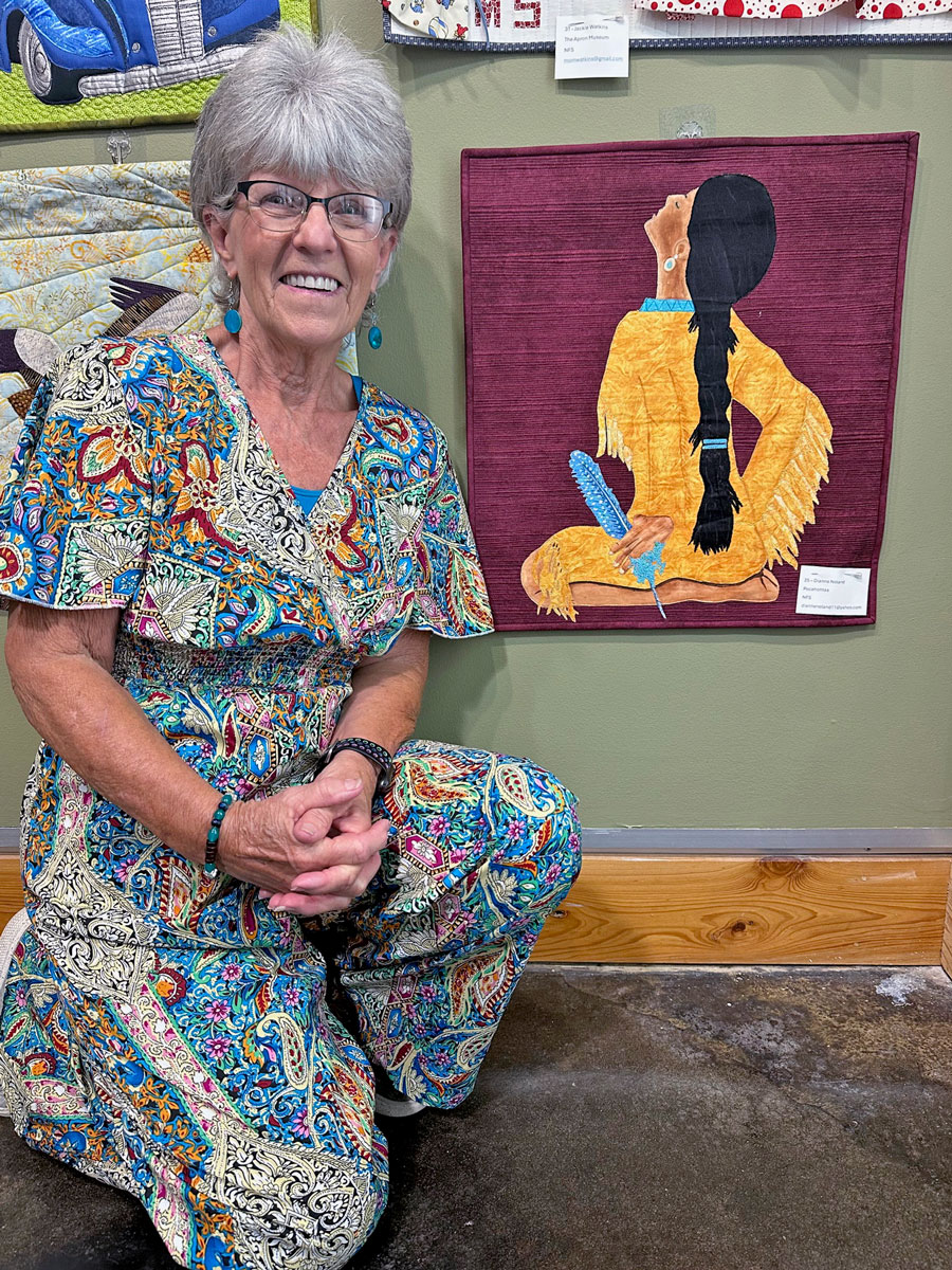 A woman kneels in front of a quilted art piece depicting a Native American