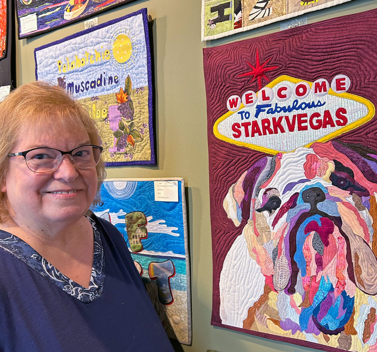 A woman stands in front of a quilted art piece depicting a bulldog and a sign that reads "Welcome to Fabulous Starkvegas"