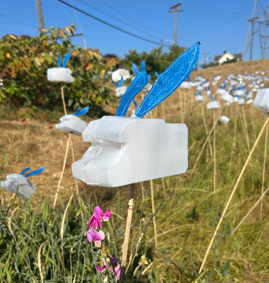 A close-up of a white, bunny-shaped object with blue ears, mounted on a thin pole in a grassy field. The bunny is made from repurposed plastic materials. In the foreground, a small pink wildflower is in focus, while more of these bunny-shaped objects can be seen scattered across the background.