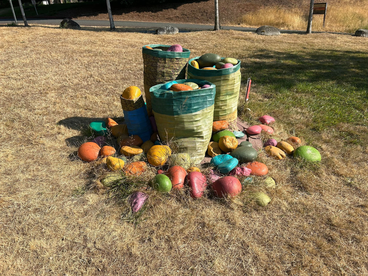 A collection of colorful, painted rocks is gathered around and inside three large, upright cylindrical baskets in a dry, grassy area. The baskets appear to be made from fabric or woven material and are weathered, blending with the surrounding environment. The rocks are painted in vibrant shades of green, orange, pink, yellow, and blue.