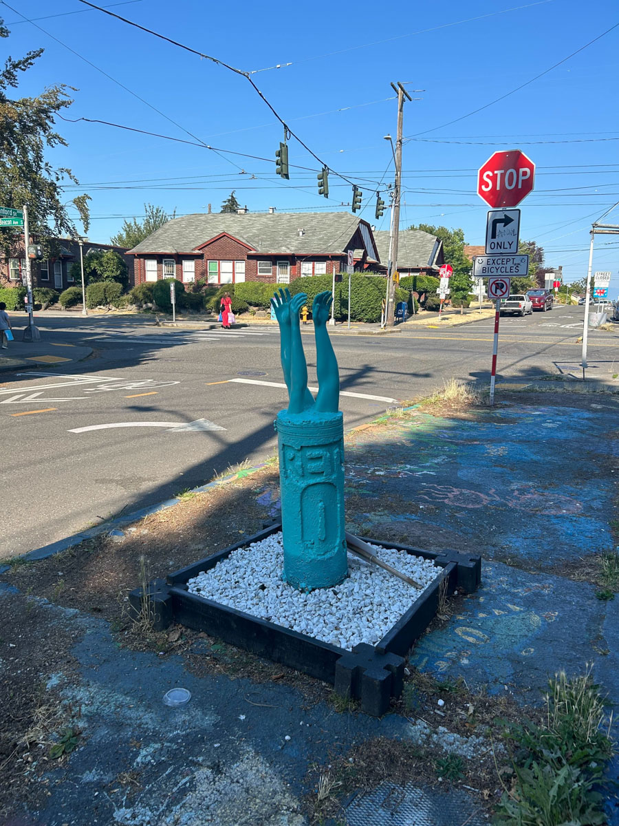 A teal-colored sculpture of a pair of human arms and hands reaching upwards is installed on a small square platform filled with white gravel at an urban street corner. The base of the sculpture resembles a fire hydrant or similar structure. The sculpture is situated near a "Stop" sign and other traffic signs, with residential houses, pedestrians, and vehicles visible in the background under a clear blue sky.