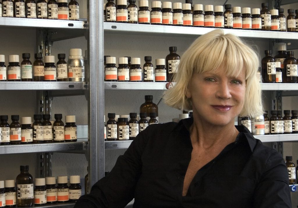 a woman with a blonde bob smiles in front of a wall of many vials