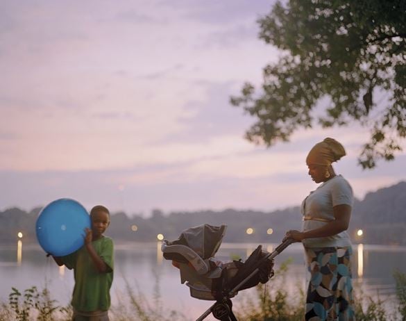 Photograph of a woman pushing a stroller with baby carrier behind a young boy holding a large blue ball all in front of a lake at dusk.