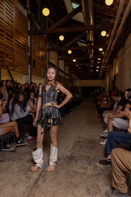 A model wears a small black dress with a gold statement belt and oversized white boots.