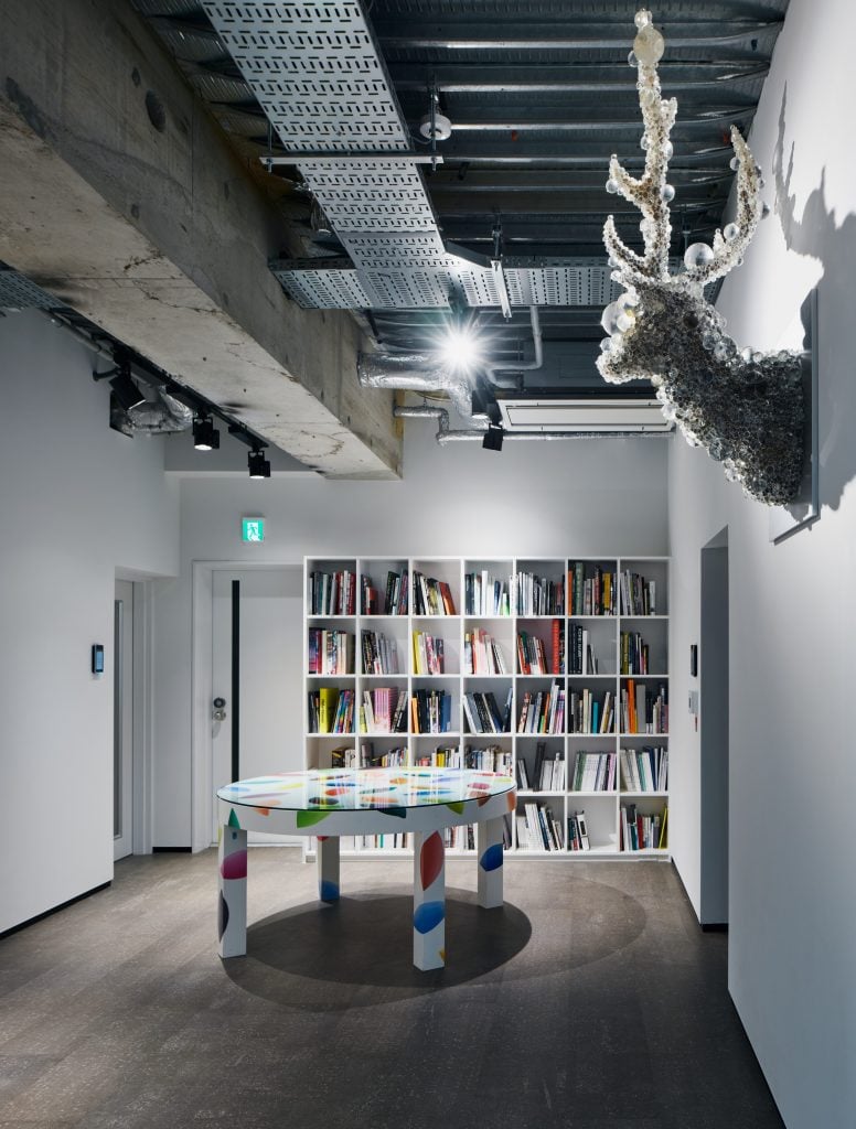 A room with a large book shelf, an artistic round table, and a sculpture of a dear covered with bubbles on the wall on the right.