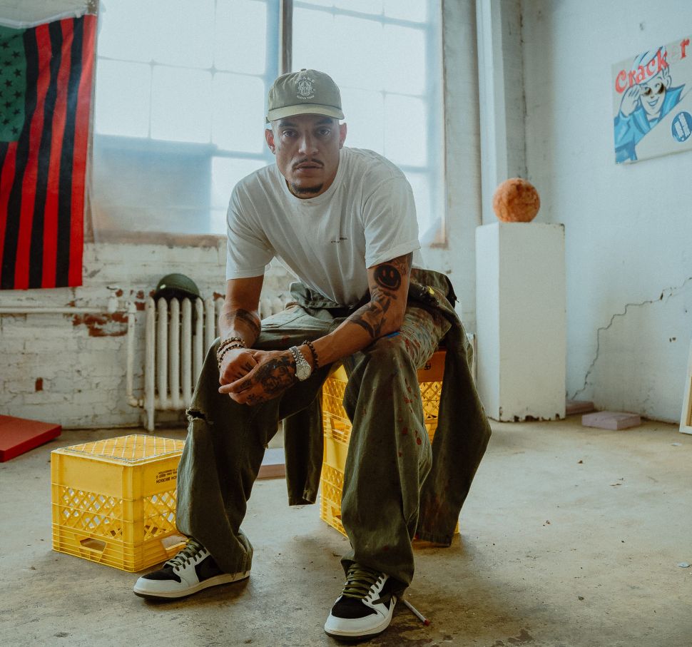 A main wearing a hat sits on a yellow milk crate in an artist's studio