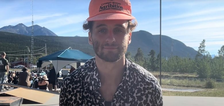 A man with a black white leopard printed button up shirt, orange baseball cap and a beard sits on a skateboard ramp