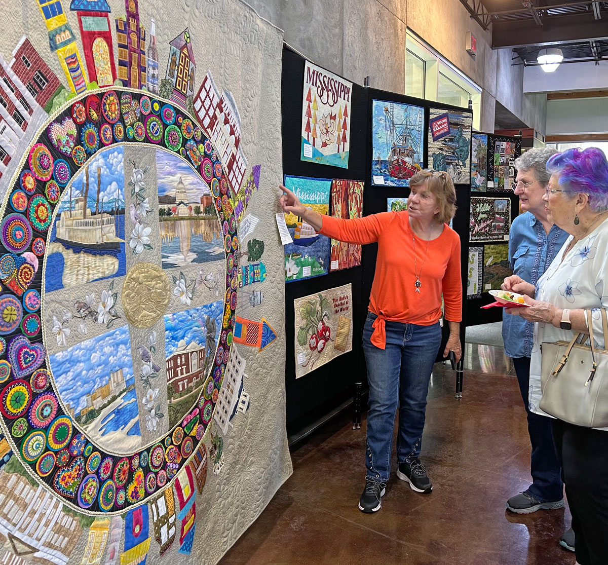 A woman points to a very large quilted art piee with a circular motif and buldings all along the edges