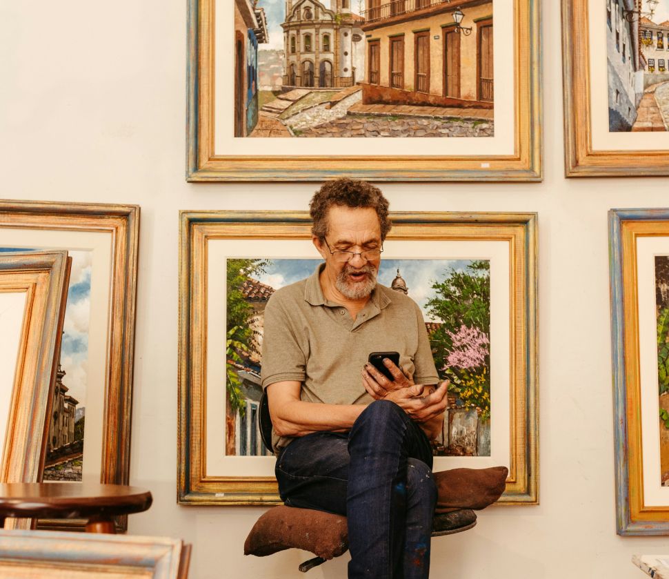 A man sits looking at a phone in front of a wall of framed artworks